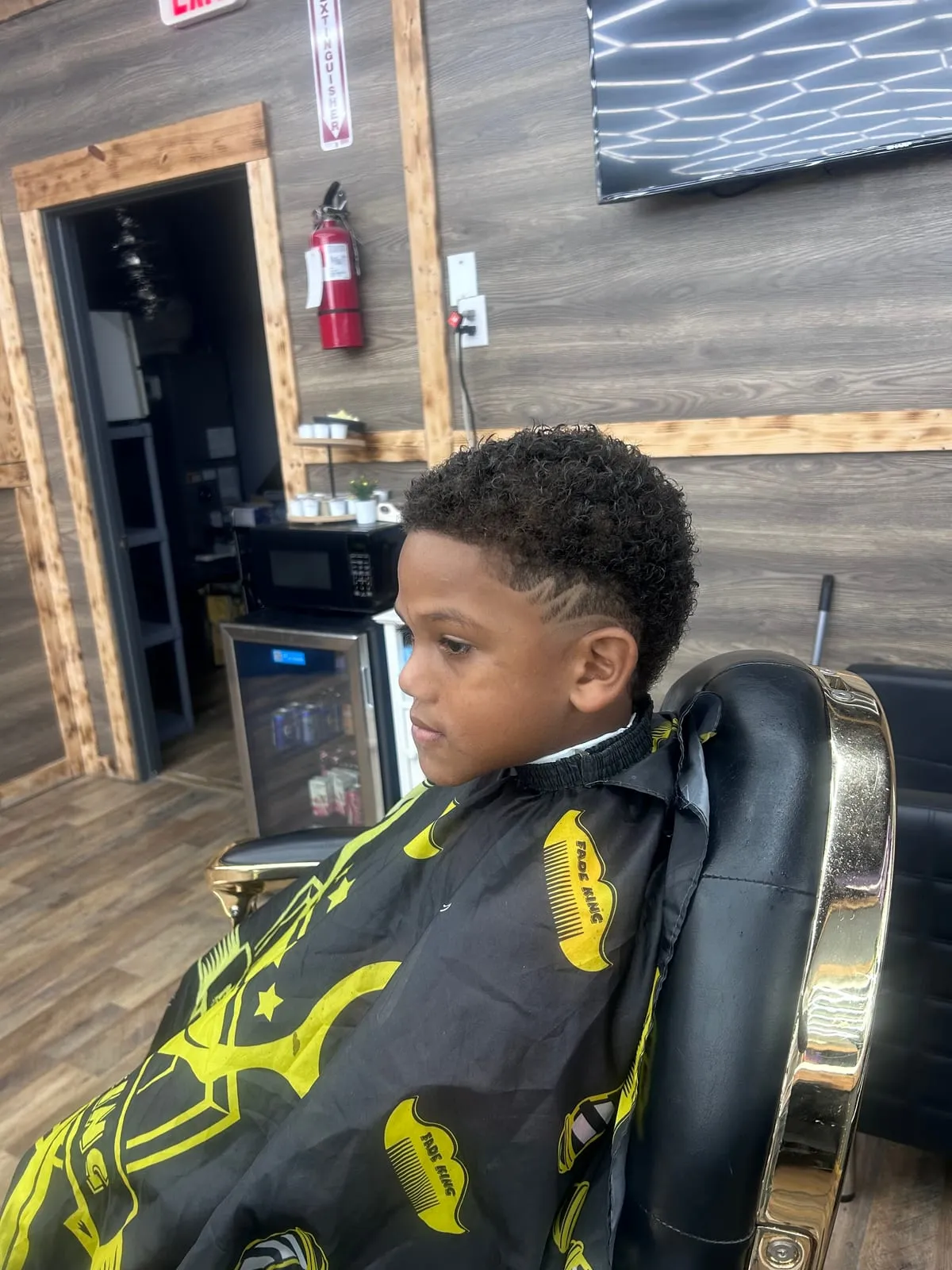 Boy sitting in a barber chair in a gold-trimmed cape, with curly hair on top and a sharp fade