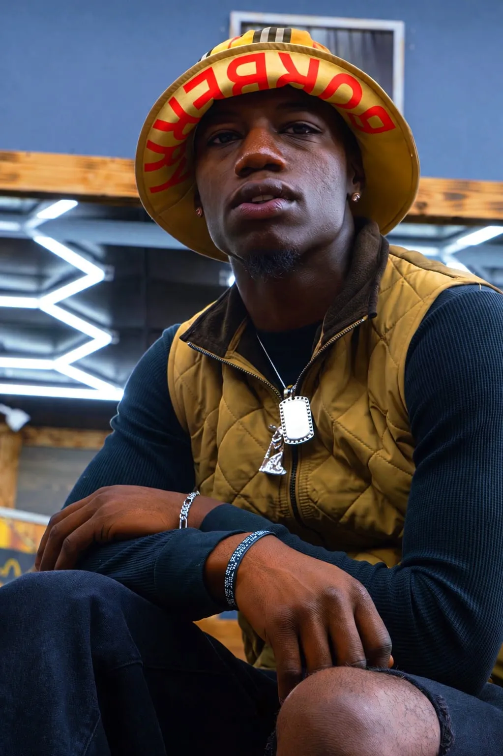 Dink, barber, seated in a yellow bucket hat and quilted gold vest under the shop's hexagon ceiling lights.