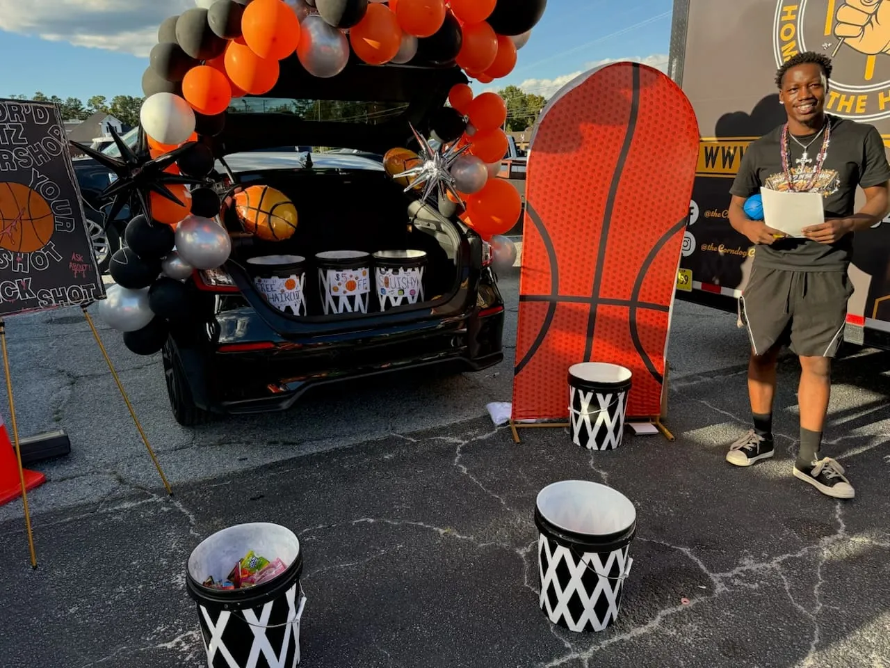 A man standing beside a car trunk decorated with orange, black and silver balloons, a basketball-hoop backboard and basketball-net buckets, in a parking lot