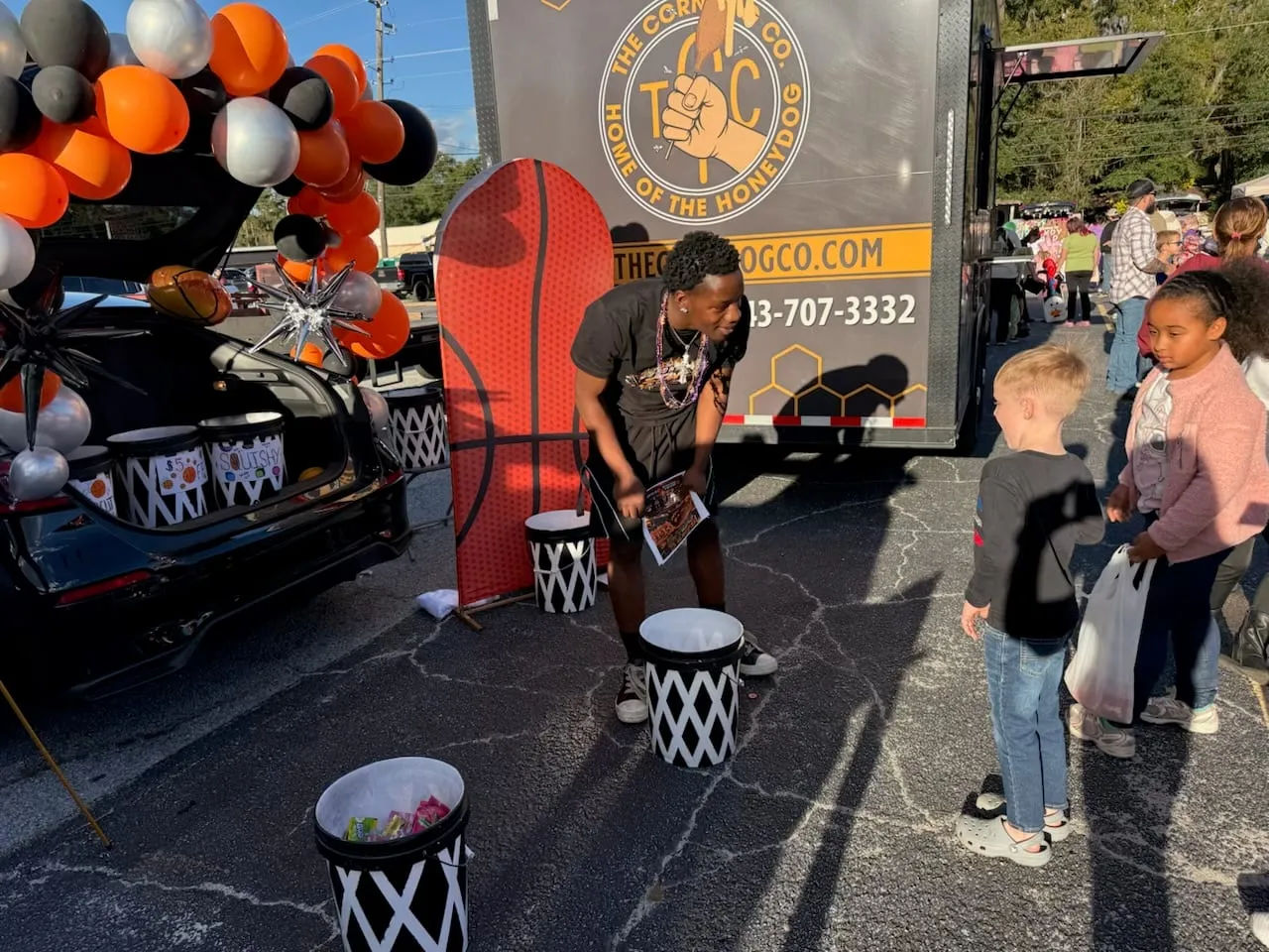 Two children talking with a man at the decorated trunk, balloon arch and basketball buckets behind them