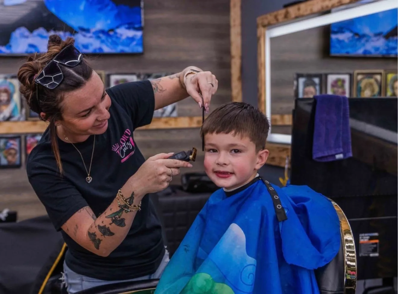 Barber trimming a young boy's hair in a blue cape in front of the shop's wood-framed mirrors