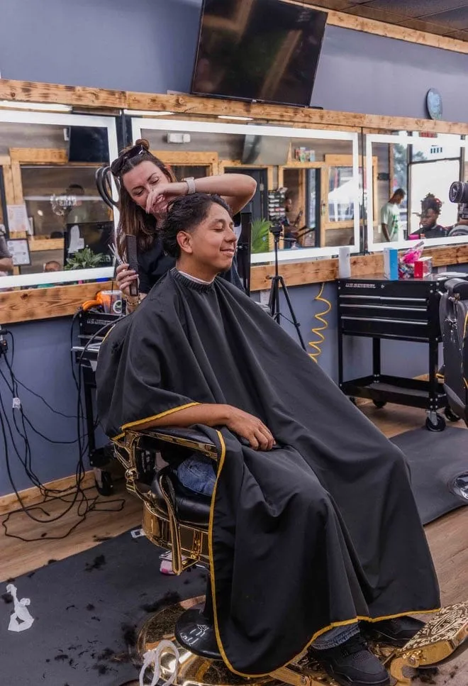 Man seated in a black-and-gold barber chair under a black cape while the barber works behind him