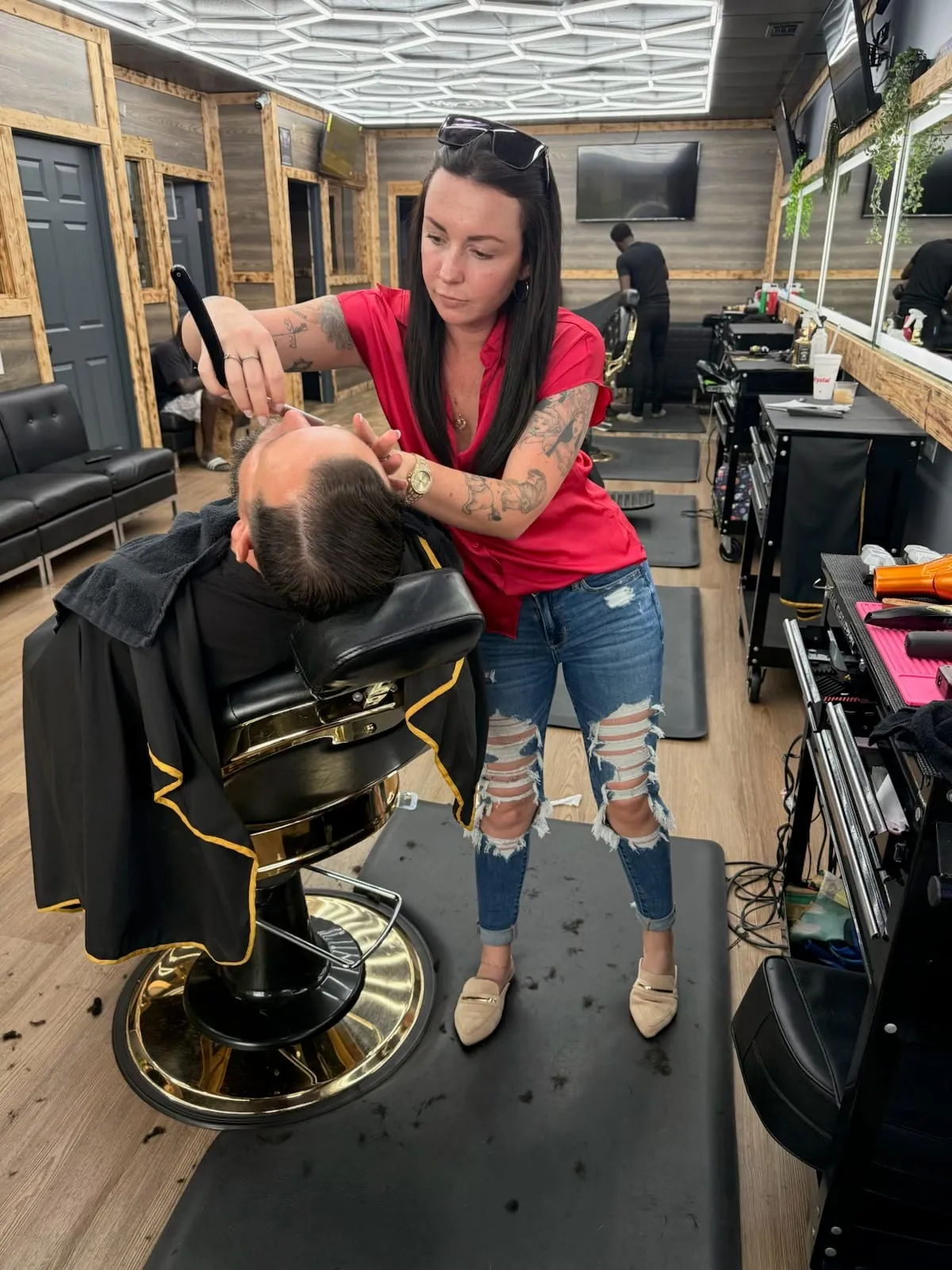 Barber in a red top working on a client who leans back in a black-and-gold chair on the shop floor