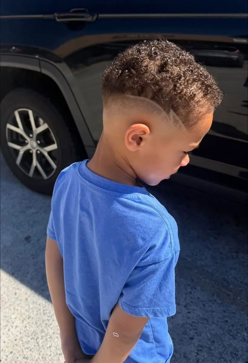 Boy seen from behind in a blue T-shirt with curly hair on top and a part shaved into the side